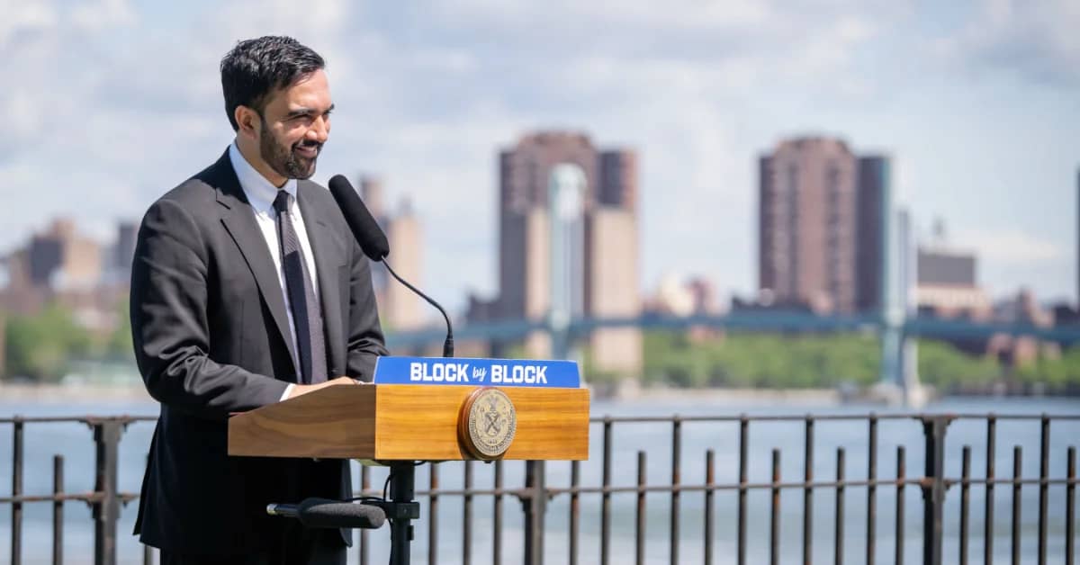 A man with dark hair and a beard wearing a black suit and tie standing at a podium with a sign that says "block by block"