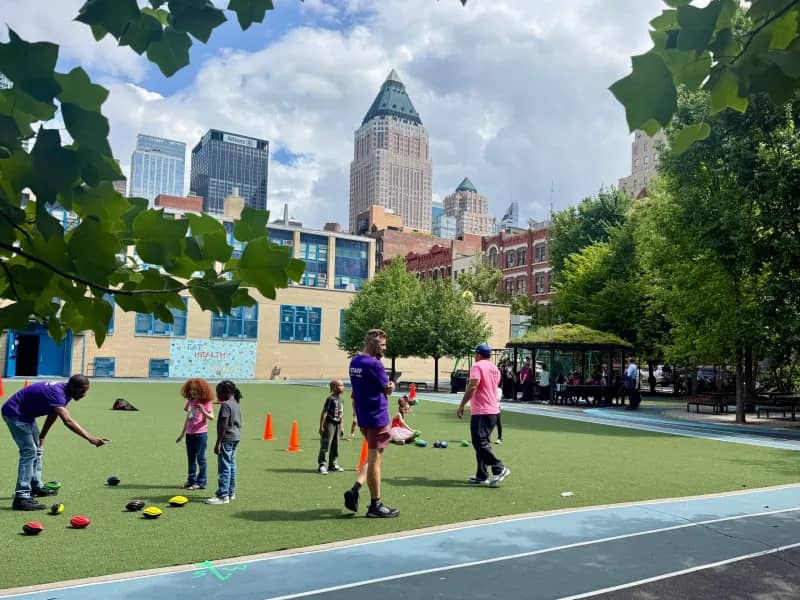 Children and adults participate in a sports activity on a synthetic grass field in an urban park, with tall buildings and skyscrapers visible in the background under a partly cloudy sky.