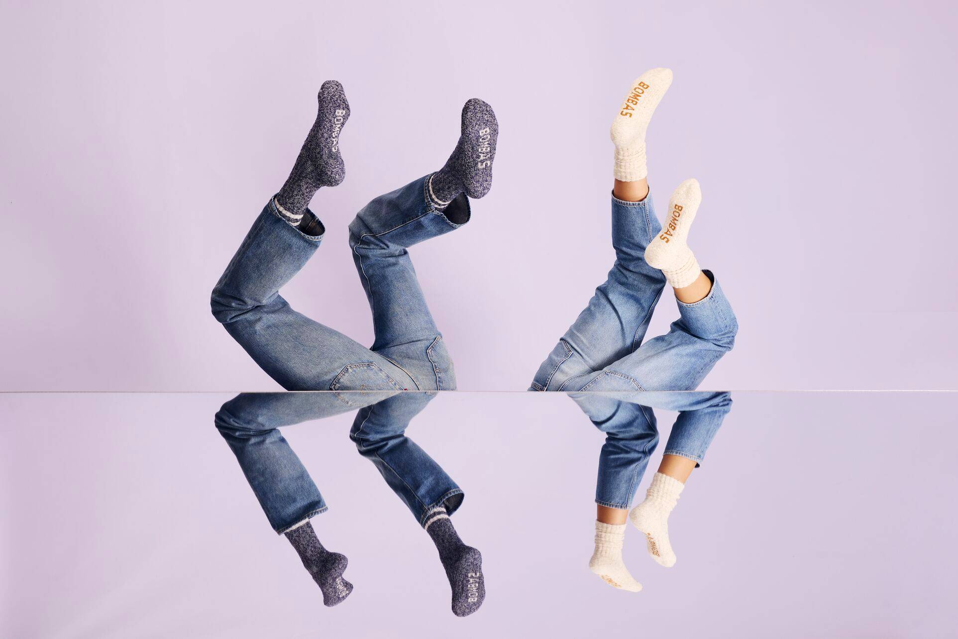 Two people with their legs up in the air, wearing jeans and bombas socks, the background is purple and their legs are reflected in a mirror in the bottom half of the image