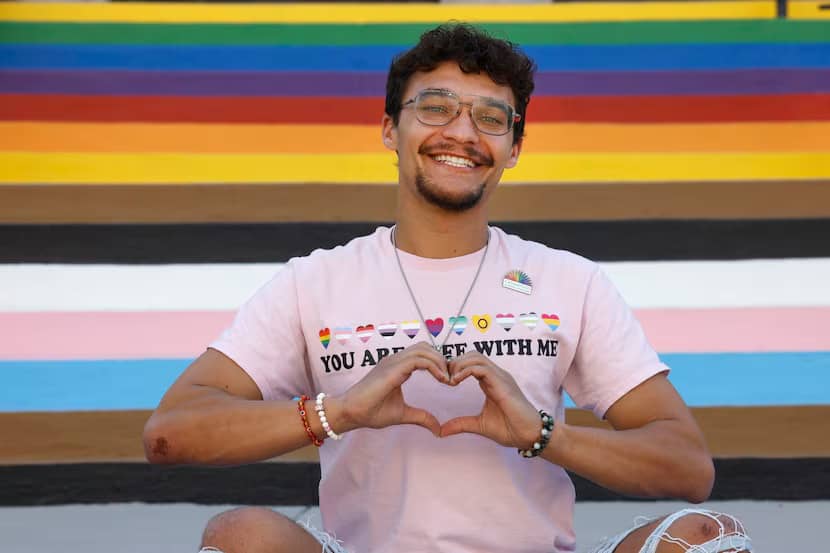 A person holding their hands in a heart, sitting and smiling in front of a rainbow-painted wall behind them