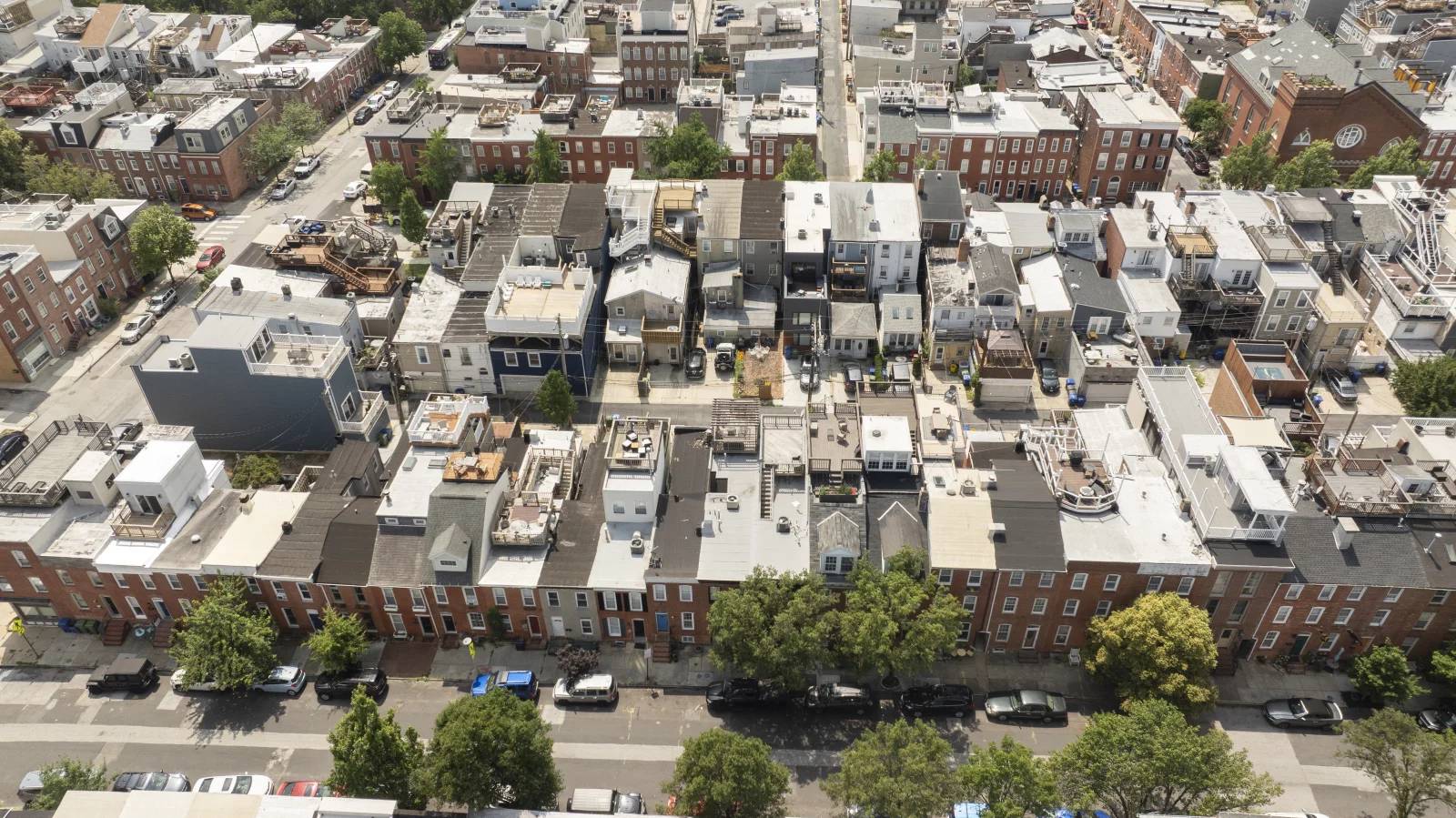 Aerial view of a dense urban residential block with rowhouses and apartment buildings, many with brick facades and flat roofs. Multiple vehicles line the street below, with construction equipment and materials visible on several rooftops. Green trees are 