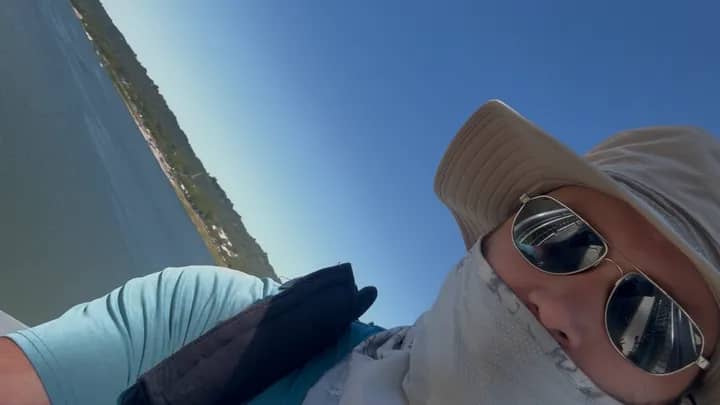 Selfie of Lee Johns wearing a hat, sunglasses, and face covering with a body of water behind him