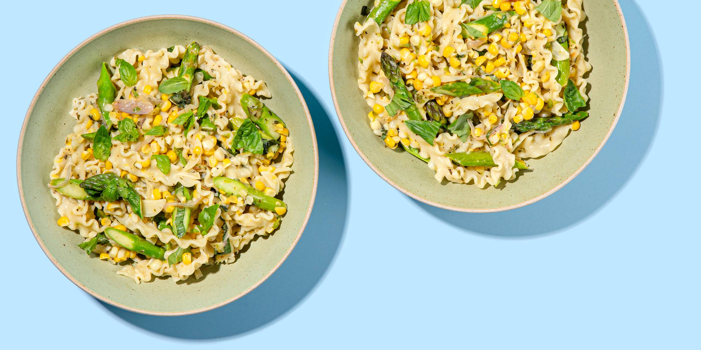 Two sage green-colored bowls filled with a pasta, corn, and greens dish on a blue background