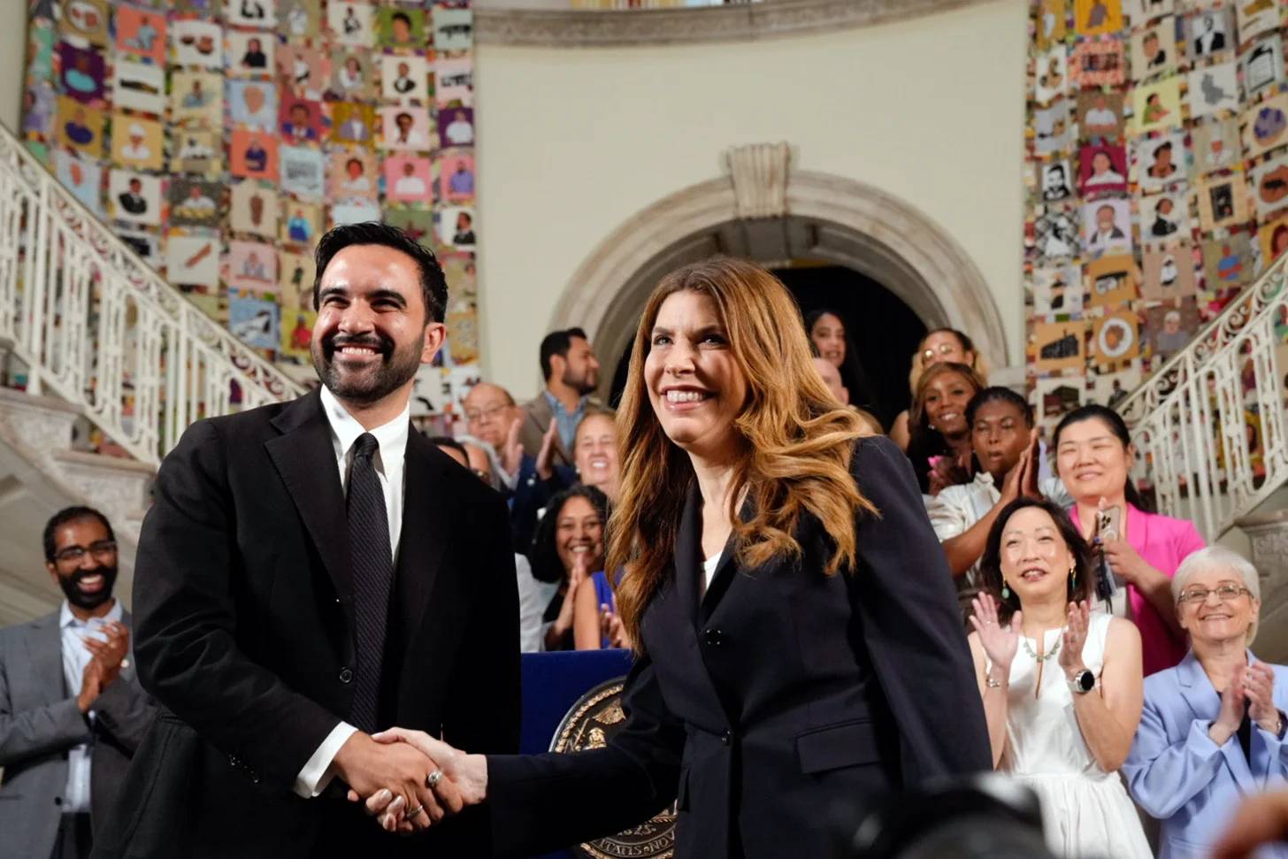 Two people in dark business suits shake hands while smiling in front of a curved doorway, surrounded by an audience of people applauding in a stairwell with colorful artwork on the walls.