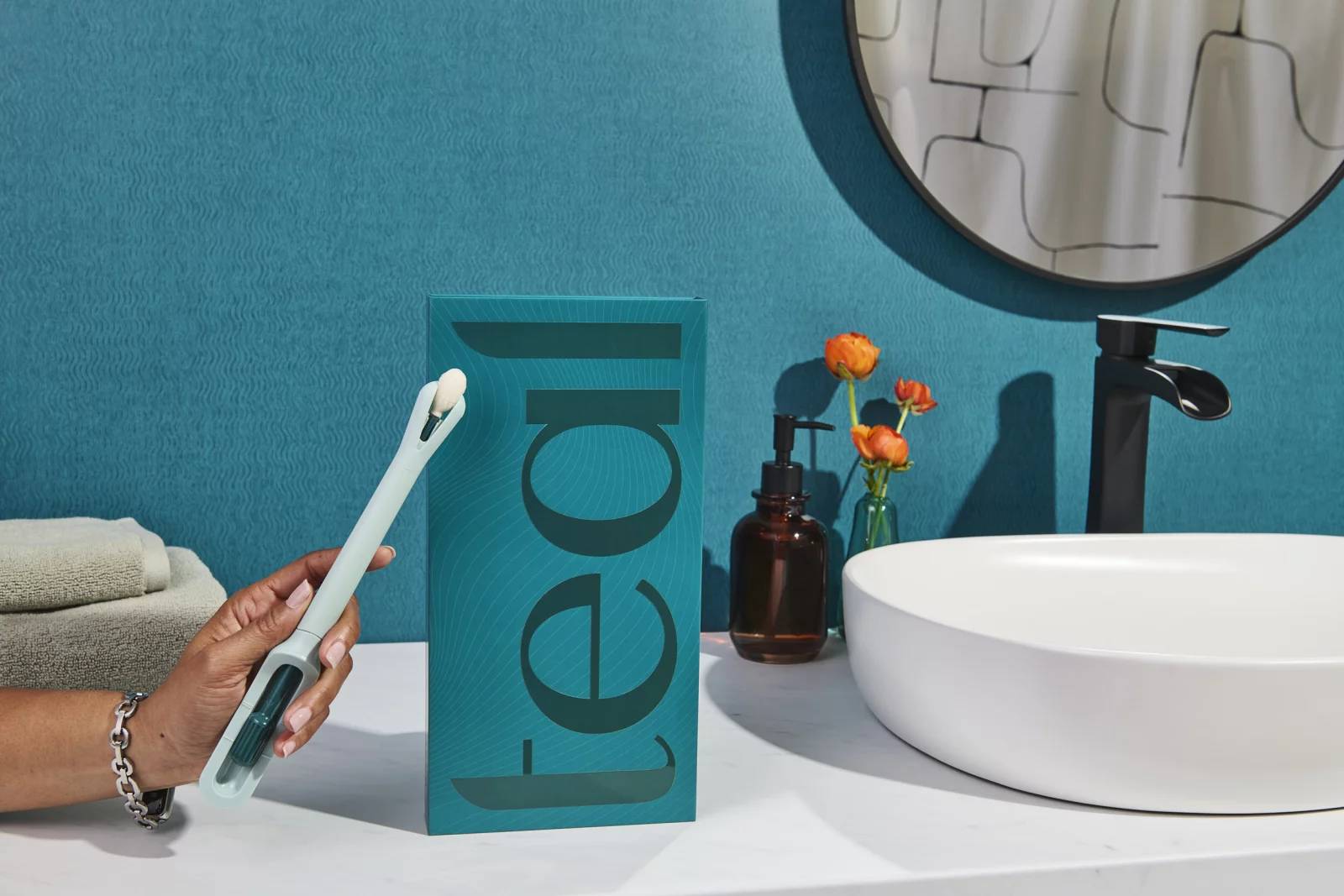 A hand coming from off-screen holding a Teal Wand next to a box on a bathroom counter