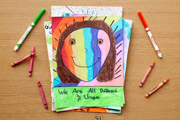 Crayons and markers next to paper with a drawing of a rainbow-colored person that reads "We are all different and unique"