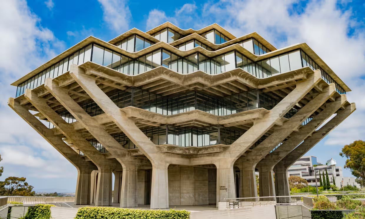 The exterior of a concrete structure with windows, the top is wider than the bottom, a blue clouded sky is in the background