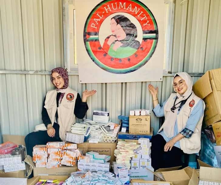 Two people in tan vests and head scarfs in front of a "Pal Humanity" banner and medical supplies