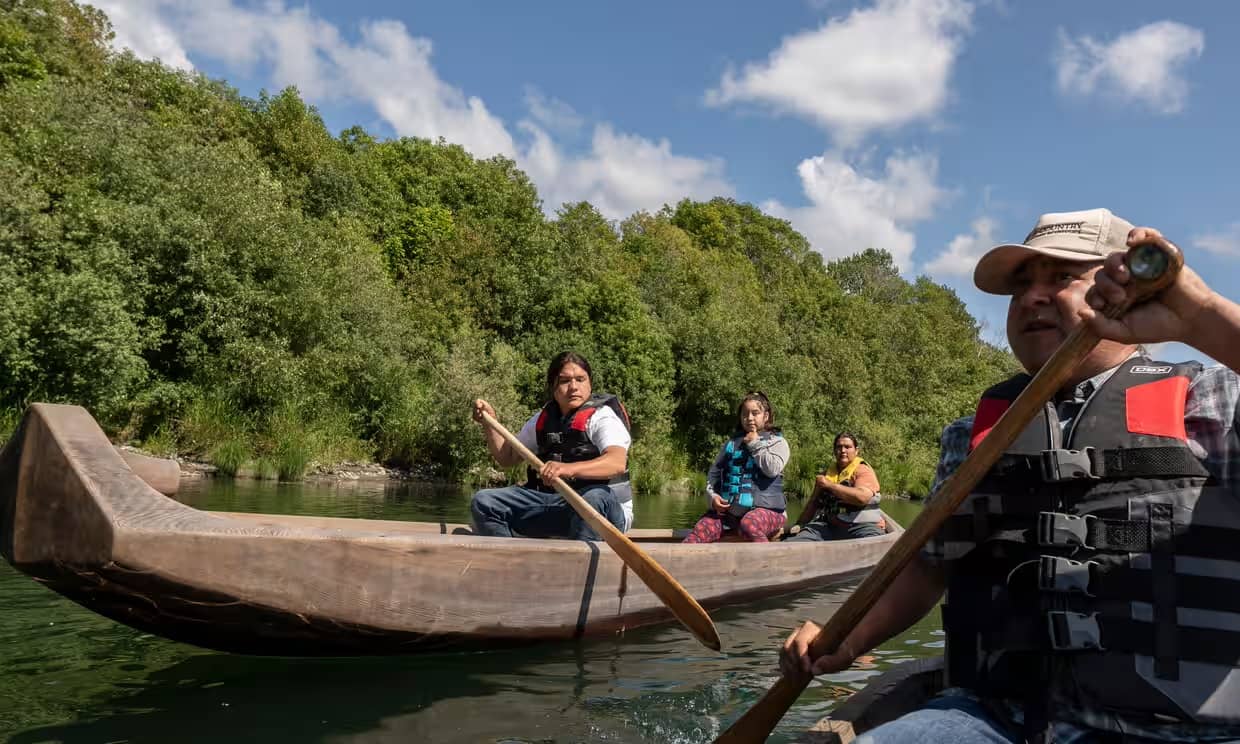 Yurok people in wooden canoe boats paddling down a waterway lined with trees