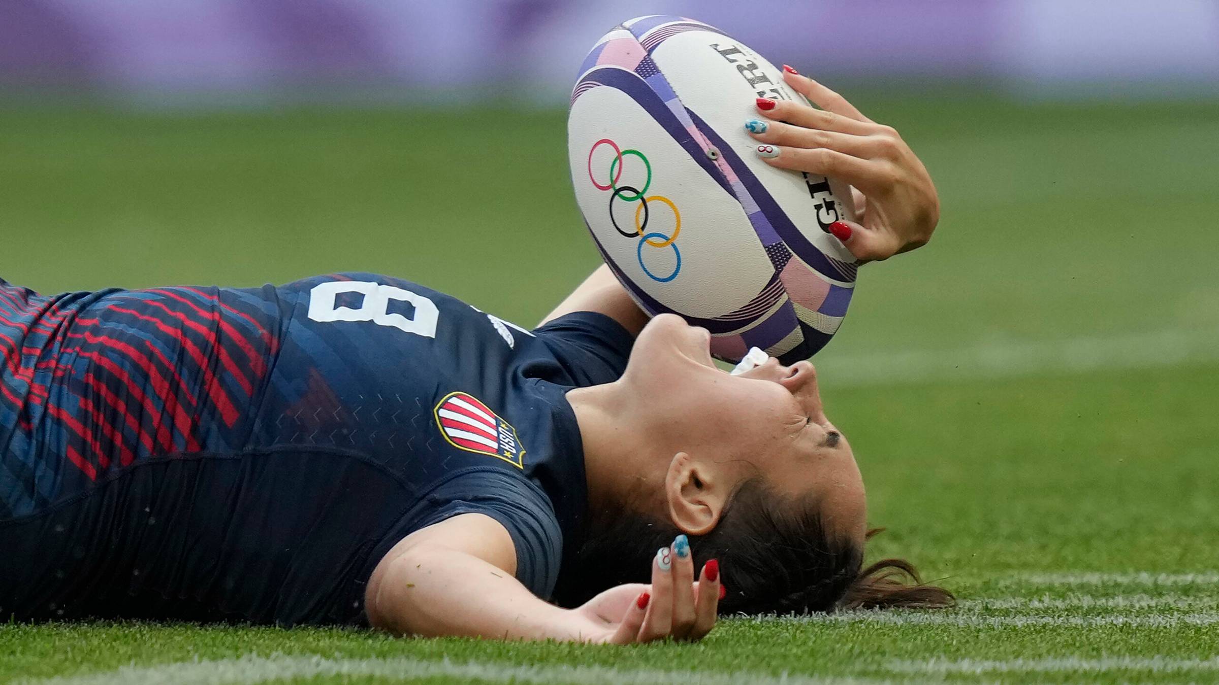 A USA rugby player lays on the ground holding a rugby ball with their mouth open in celebration