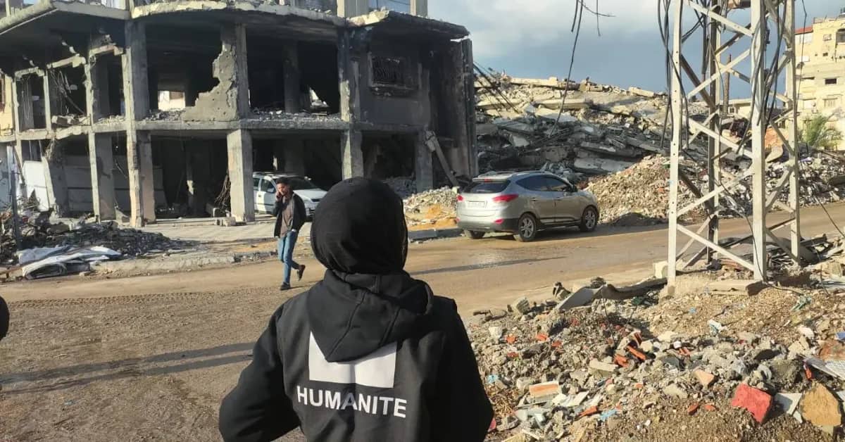 Person wearing a black jacket labeled "HUMANITE" stands in foreground overlooking a devastated urban area with destroyed concrete buildings, rubble, and parked vehicles.