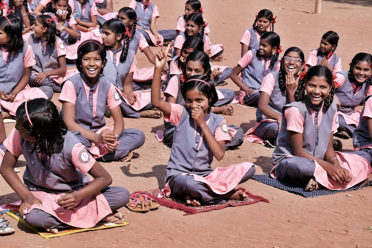 Large group of girls in pink and gray school uniforms sitting on the ground, many smiling and laughing, in an outdoor setting with brown earth.