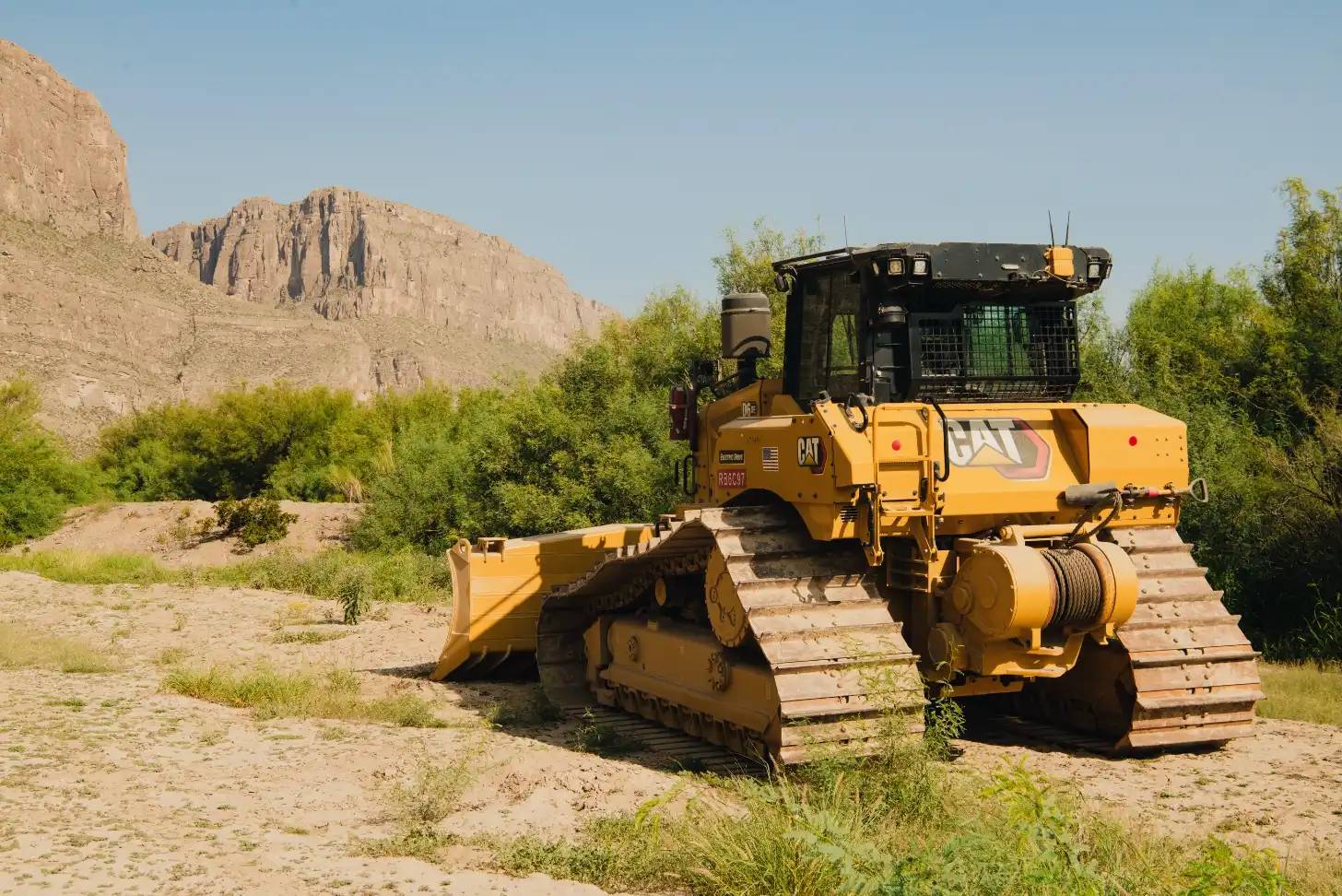 Yellow Caterpillar bulldozer with blade and roller attachment positioned on sandy ground with green shrubs and a rocky cliff formation in the background under clear sky.