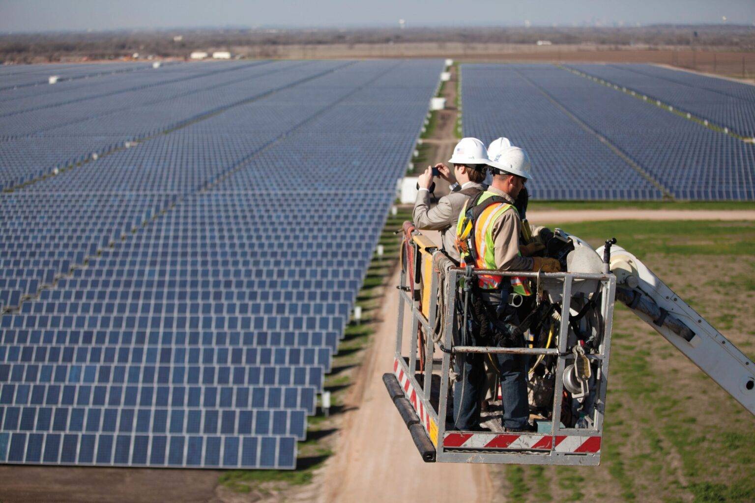 Two workers in hard hats and safety vests stand in a lift platform, inspecting vast rows of solar panels extending across a flat landscape.