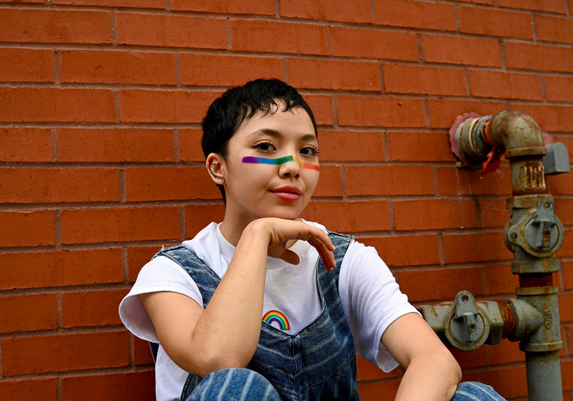 A young person with short black hair wearing a white t-shirt and denim vest sits against a red brick wall, resting their chin on their hand. They have rainbow-colored face paint across their eyes. Metal pipes and fixtures are visible on the brick wall to their right.