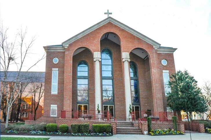 Exterior of a red-brick church with a white cross on top of it, four columns in the front