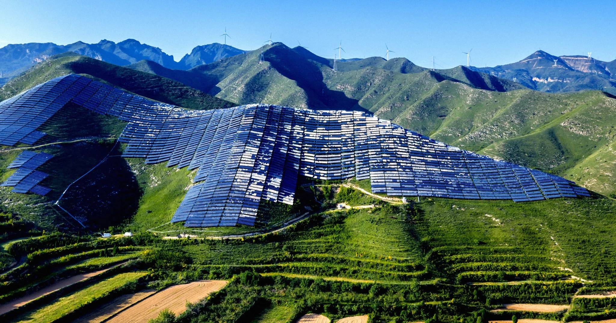 Large-scale solar panel arrays installed on hillsides in a mountainous, forested region, with wind turbines visible on distant peaks under clear blue sky.