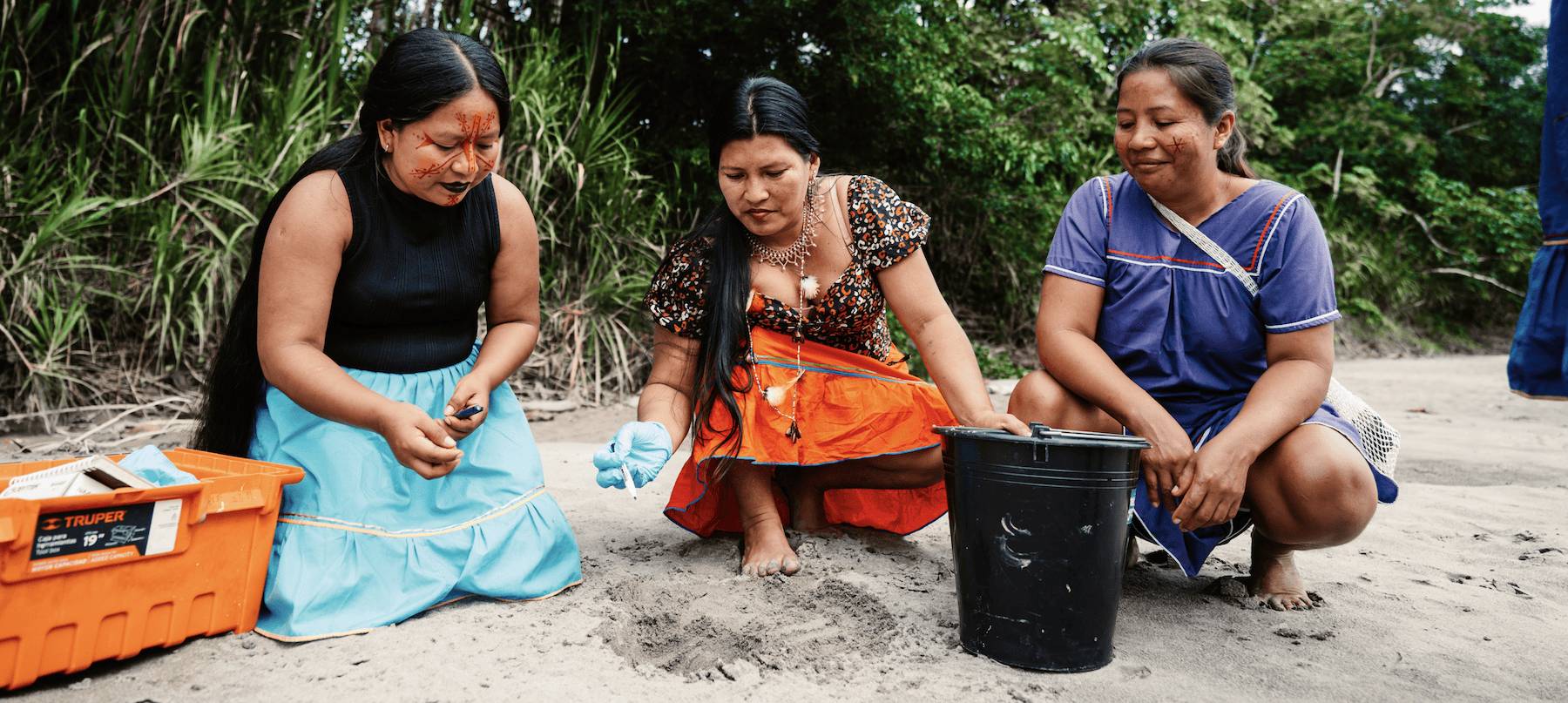 Three women in colorful dresses and skirts crouching in the sand next to an orange bucket and a black bucket.