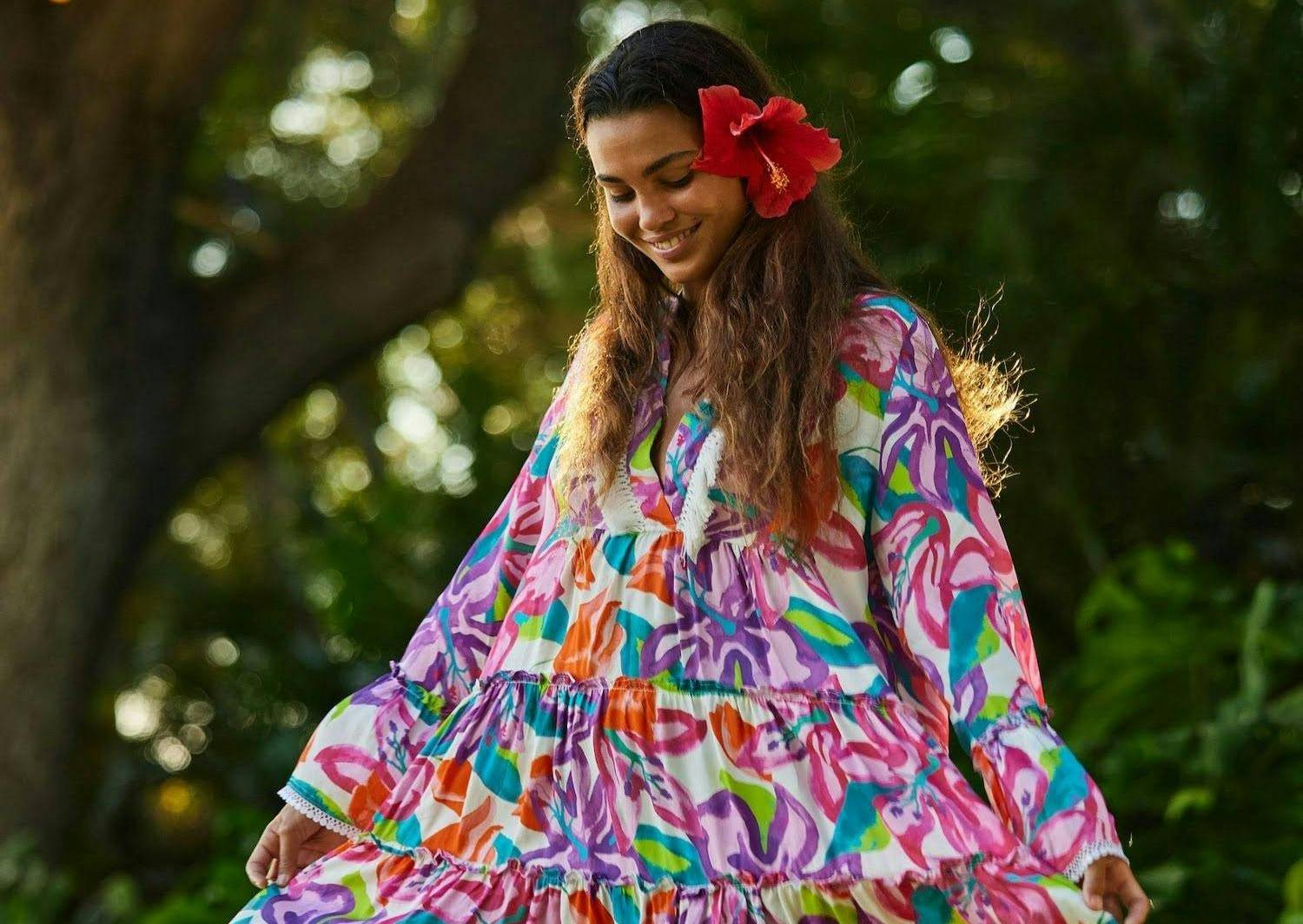 A woman wearing a colorful, long-sleeved dress with a hibiscus flower in her ear twirling underneath a large tree
