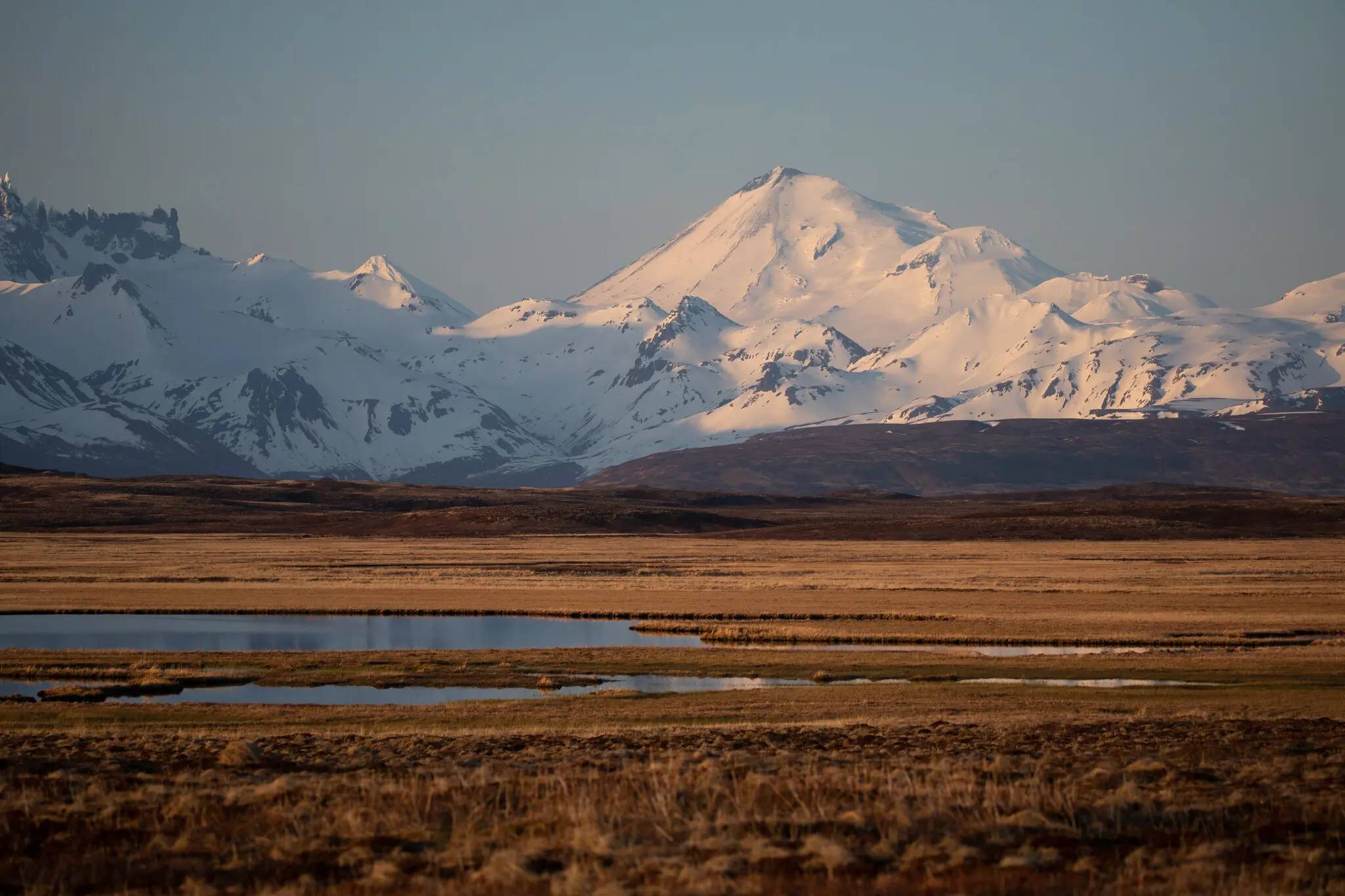 Snow-covered mountain peak rises above a broad tundra valley with scattered ponds and brown vegetation under clear sky.