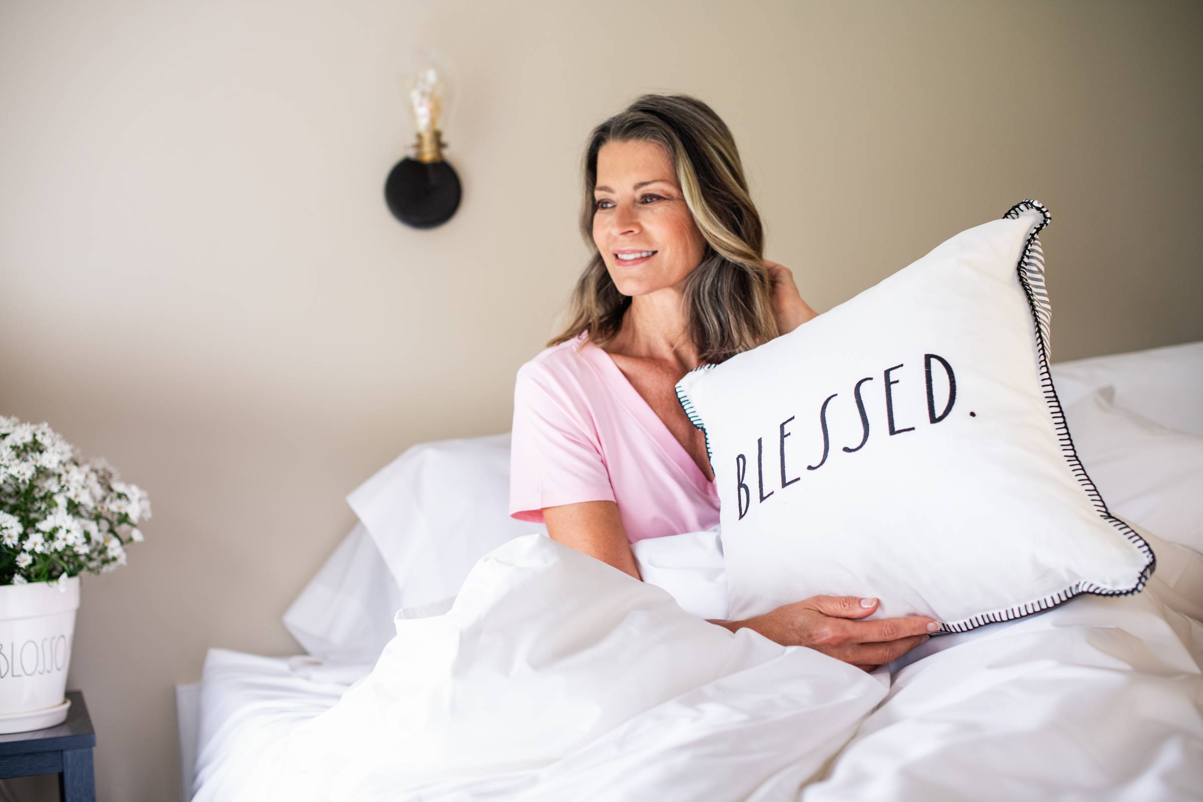 A person in pink pajamas holding a throw pillow that says "Blessed." sitting in a bed made with crisp white linens