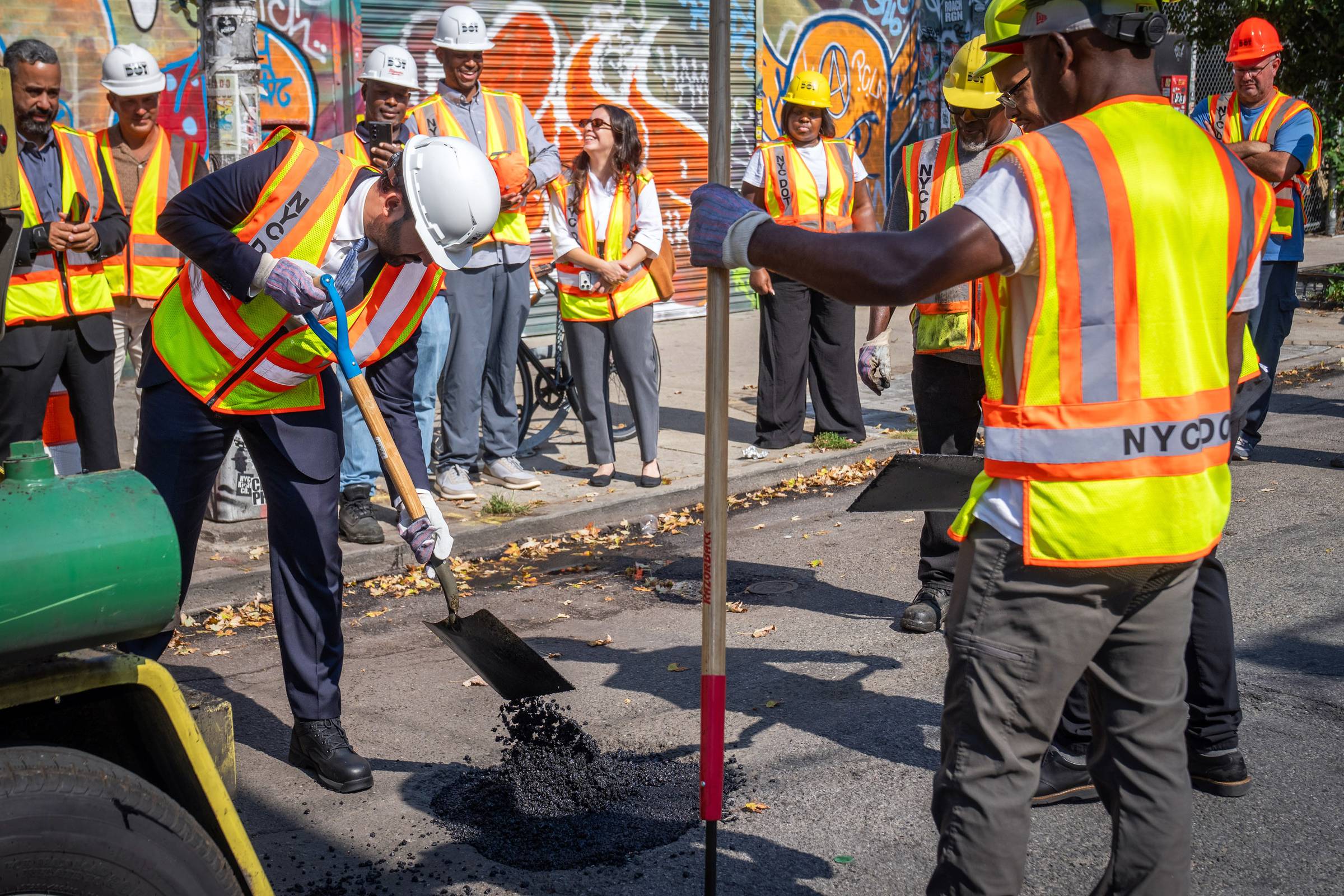 A construction worker in a white hard hat and yellow safety vest shovels asphalt during a groundbreaking ceremony. A group of workers and officials in safety gear stand watching on a city street with colorful graffiti visible on a metal shutter behind them.
