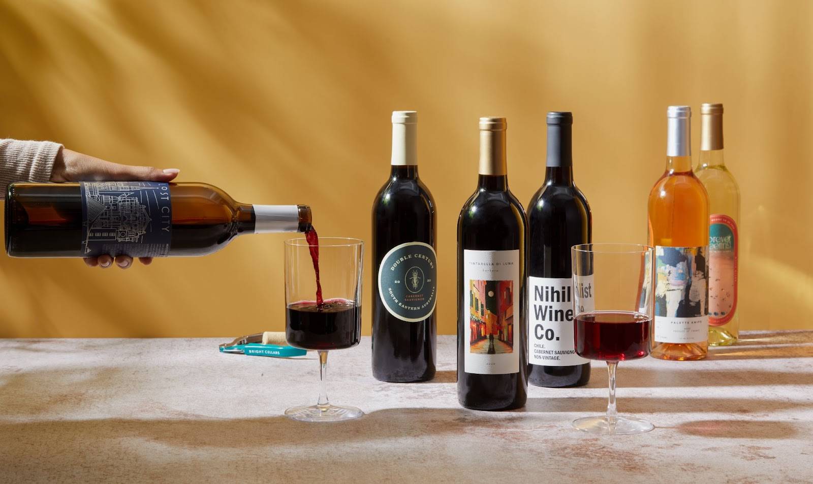 Five bottles of different kinds of wine sitting on a concrete countertop against a shadowed yellow background, and a hand is pouring from one bottle into a glass.