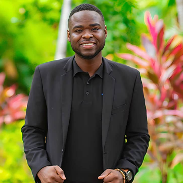 Rayvon Steward in a black polo and blazer smiling at the camera with tropical foliage in the background