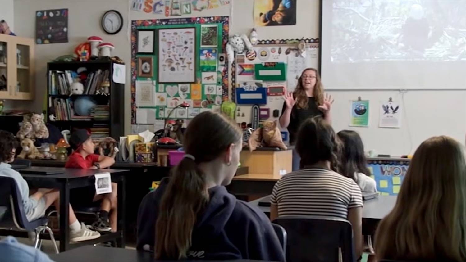 A teacher at the front of class full of students, a projection screen behind her has a bald eagle nest on it