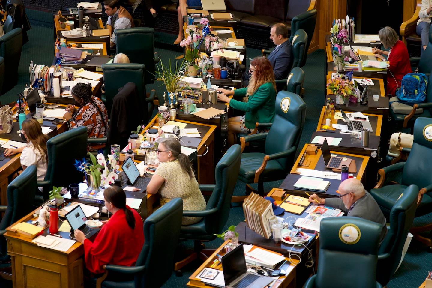 Overhead view of a parliamentary chamber with rows of green leather chairs at wooden desks. Multiple legislators, mostly women, sit at their desks with papers, laptops, and flowers. Some stand or converse while others work at their stations.