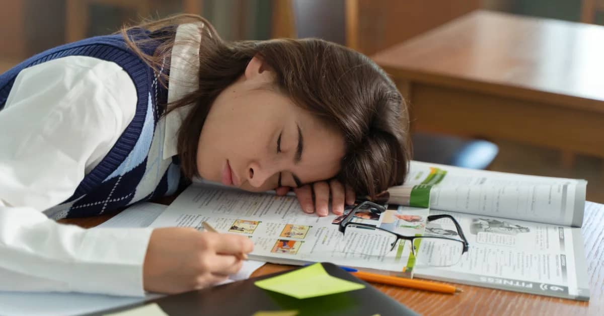 A child rests their head on an open newspaper at a wooden desk, eyes closed, with glasses and a pencil nearby and a yellow sticky note in front of them.