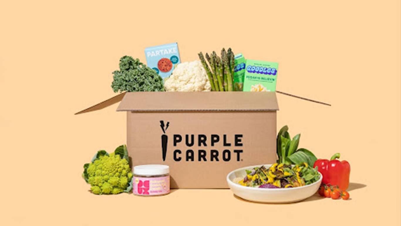 A brown Purple Carrot branded box with vegetables coming out of the top and other food products around it