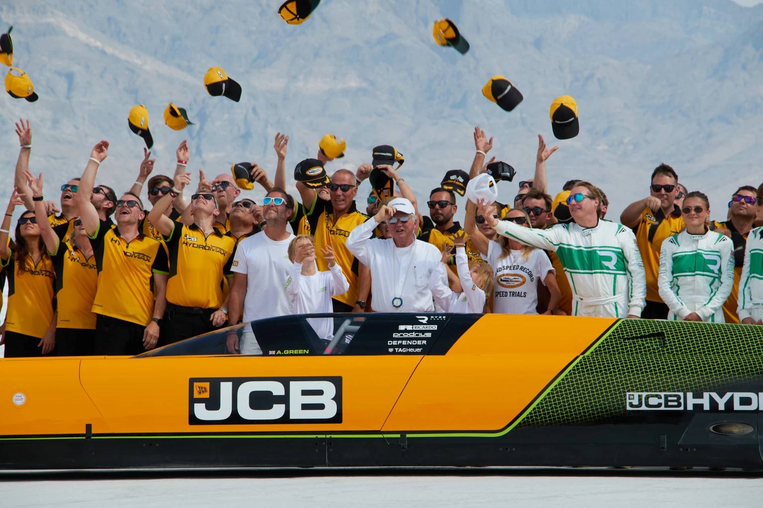 A large group of people in yellow and white clothing stand on a JCB-branded vehicle with their arms raised, throwing yellow and black caps into the air against a blue sky and mountainous background.