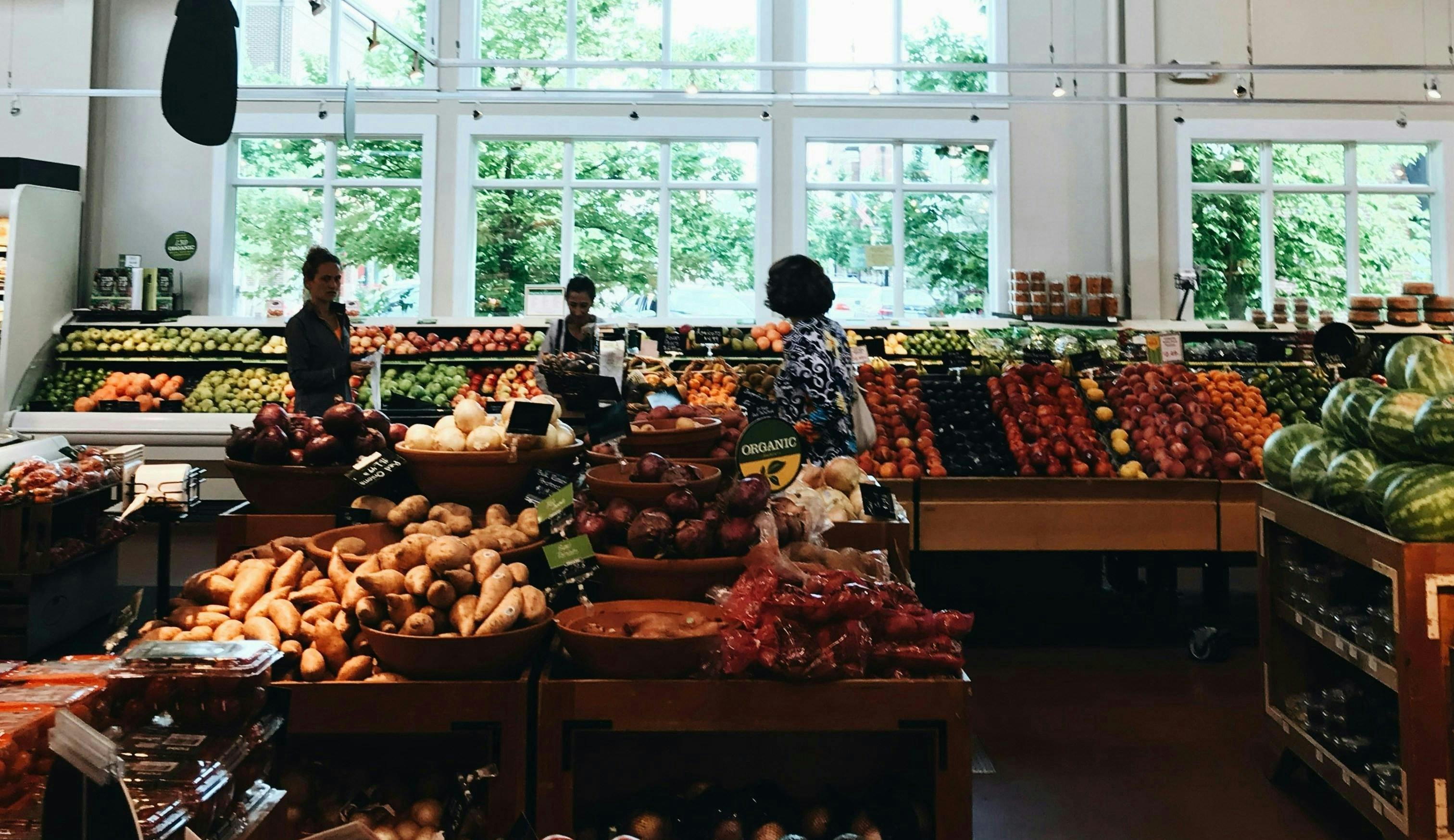 Produce section of a grocery store, there are three people shopping and the back wall is filled with windows