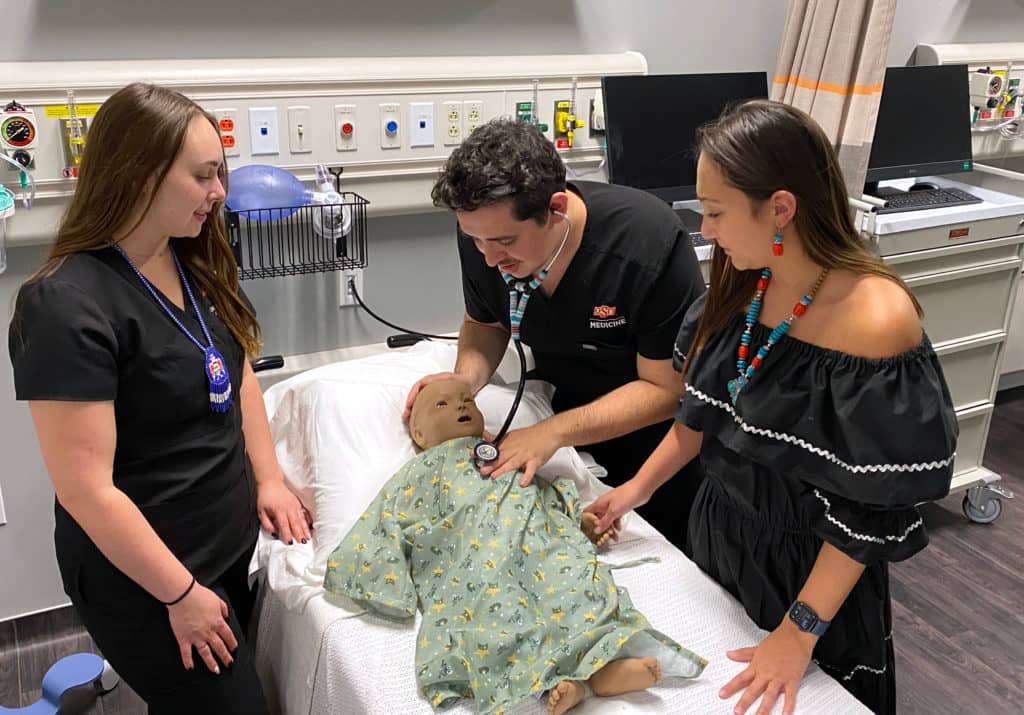 Three doctors practice using a stethoscope on a dummy patient