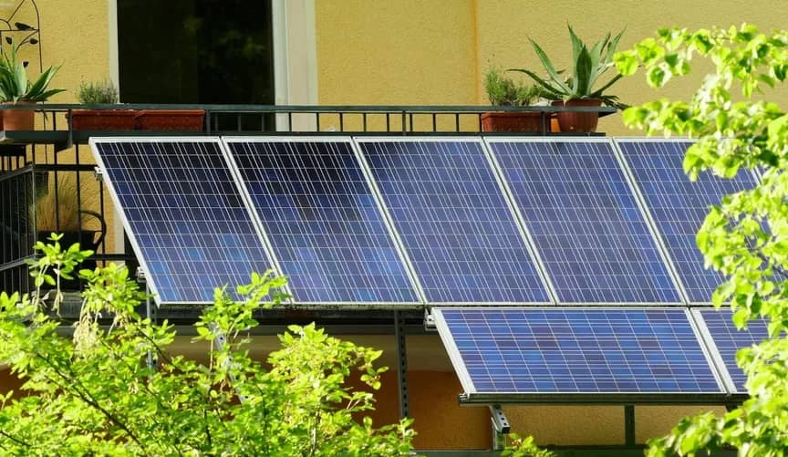 Multiple blue solar panels mounted on a metal frame against a yellow building facade, with green foliage visible in the foreground and potted plants on a railing above.