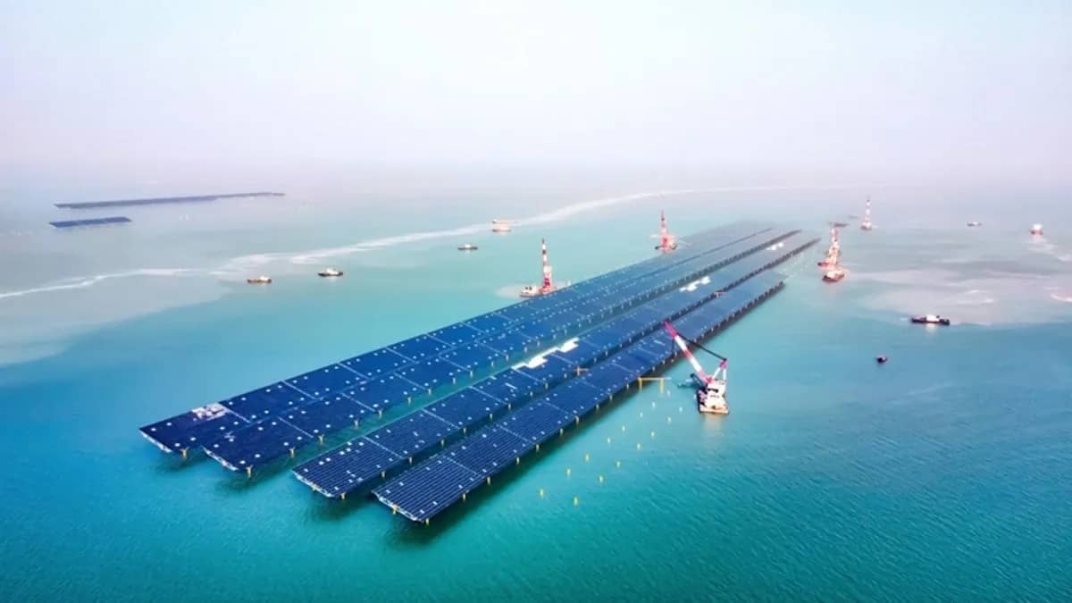 Four rows of solar panels sitting in a body of teal-blue water, there are construction cranes around the panels