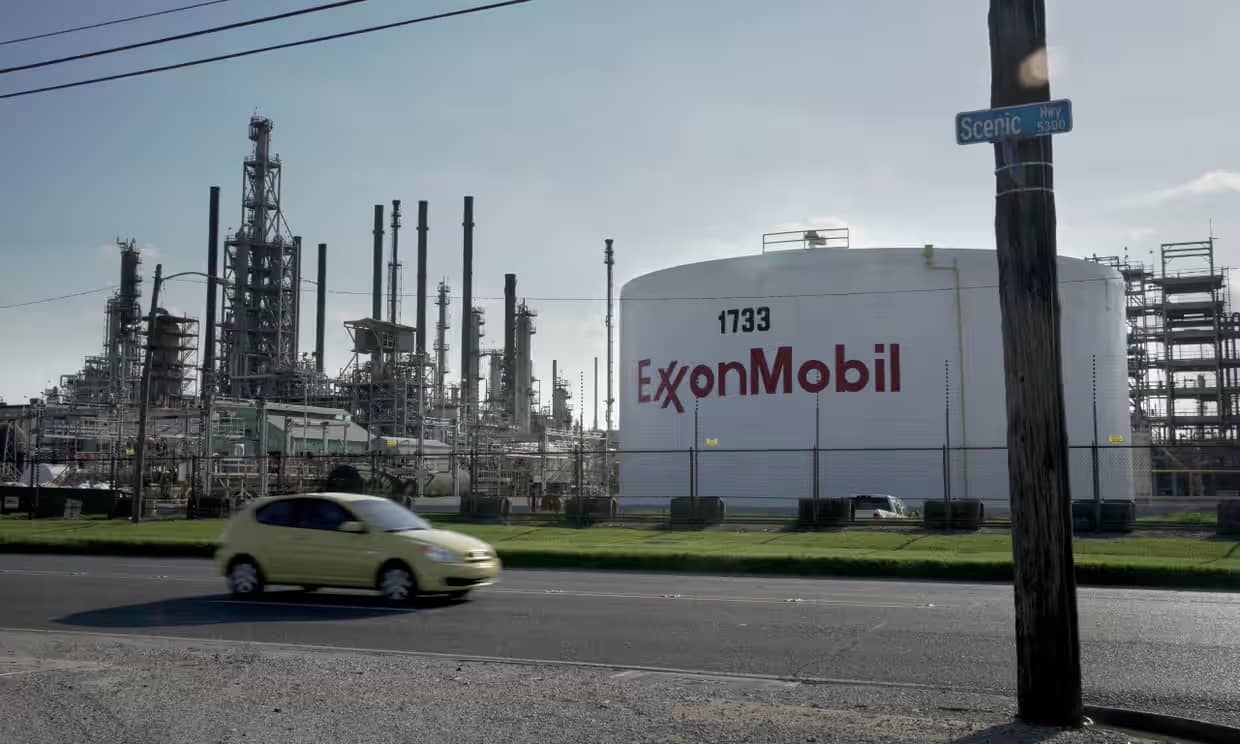 A small yellow car driving past a large white tower with "ExxonMobiile" on the exterior in red