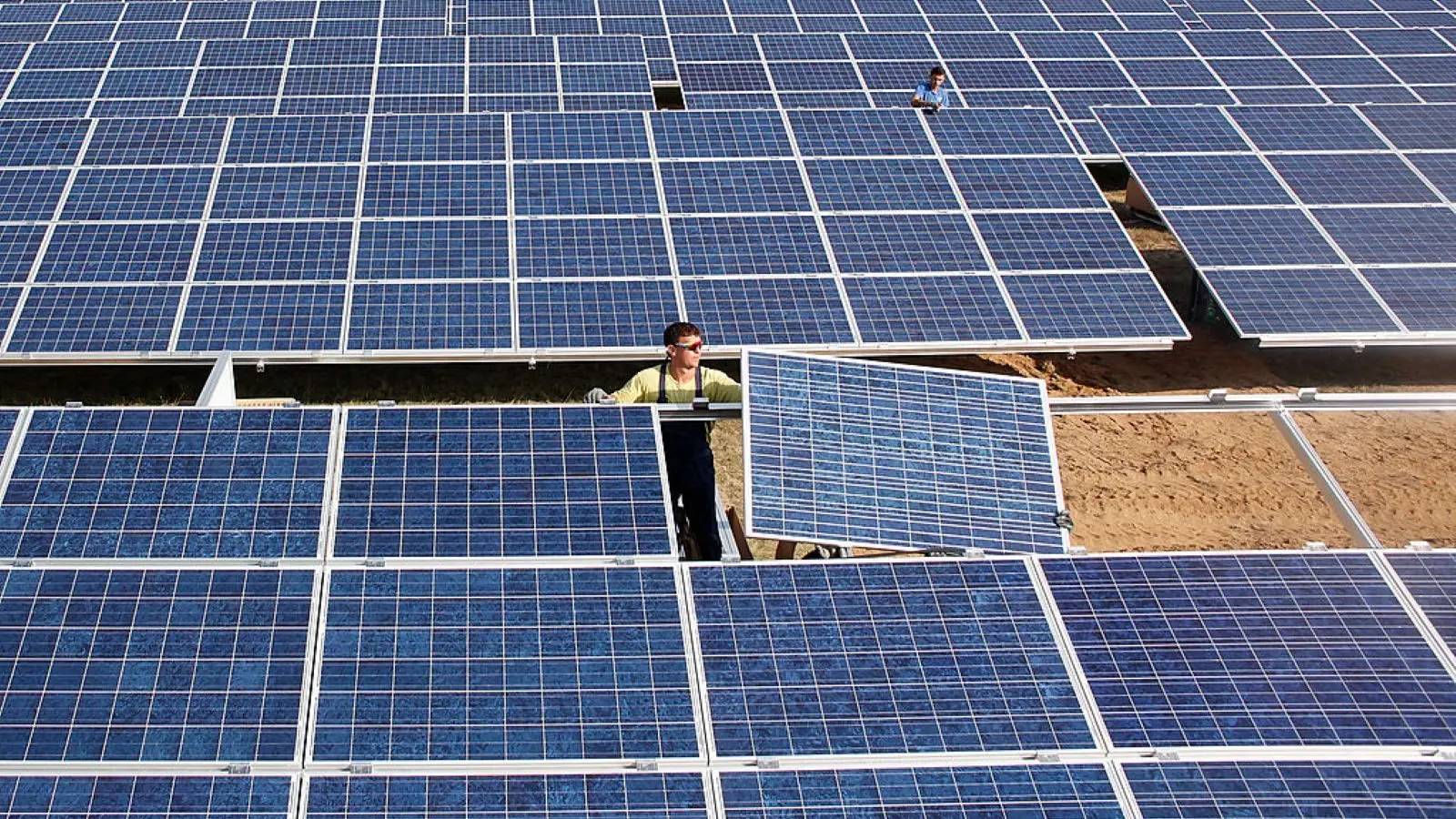 Workers walk among rows of solar panels at a large photovoltaic installation. Multiple people are visible at different points across the blue-paneled array in an open, sandy area.