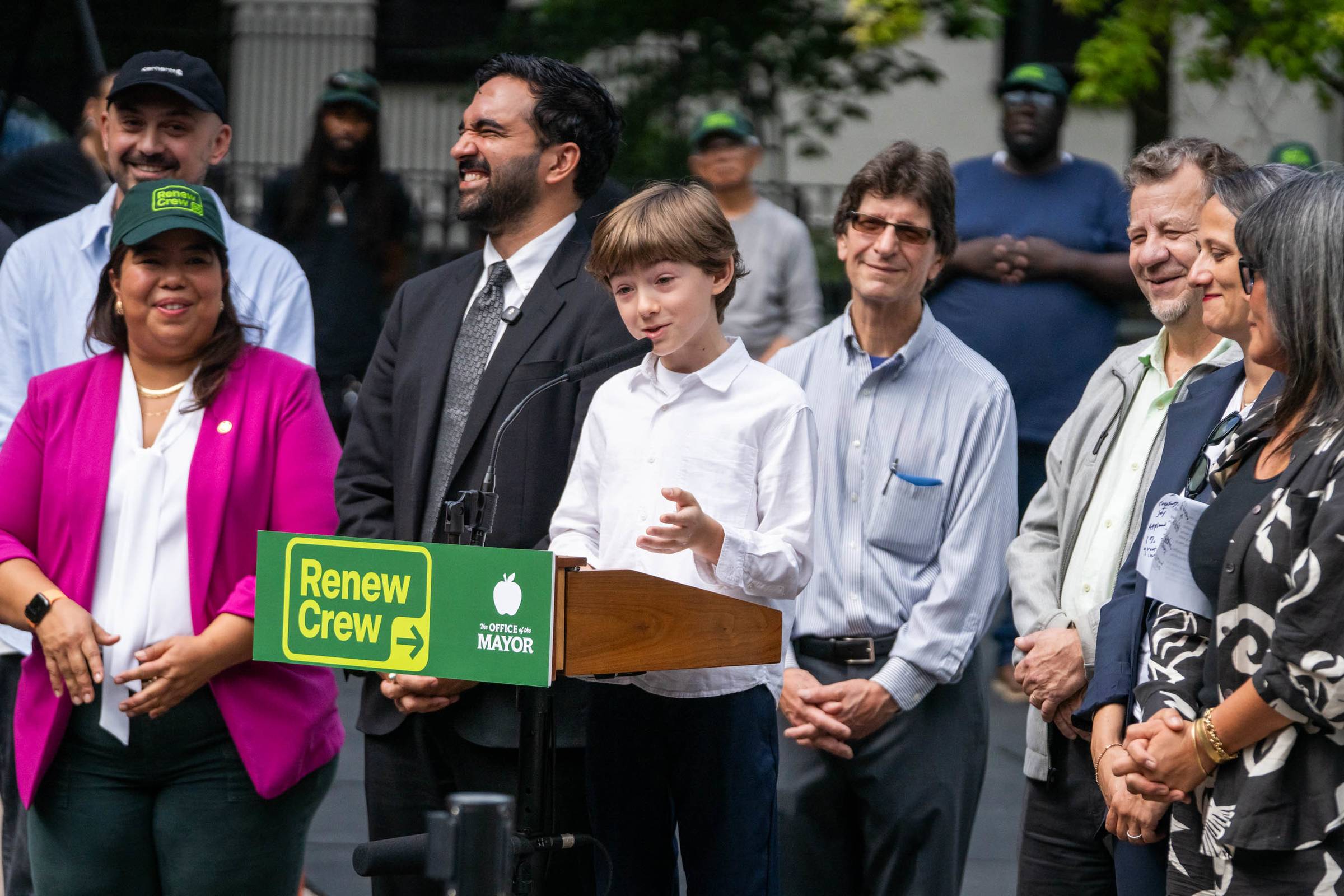 A young person stands at a podium with a "Renew Crew" sign and "The Office of the Mayor" logo, speaking to a group of officials and staff members gathered outdoors.