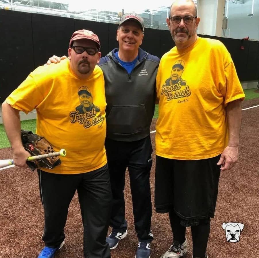 Tony Vlahovic standing in the middle of two athletes wearing yellow shirts and black bottoms, one his holding a baseball bat and glove