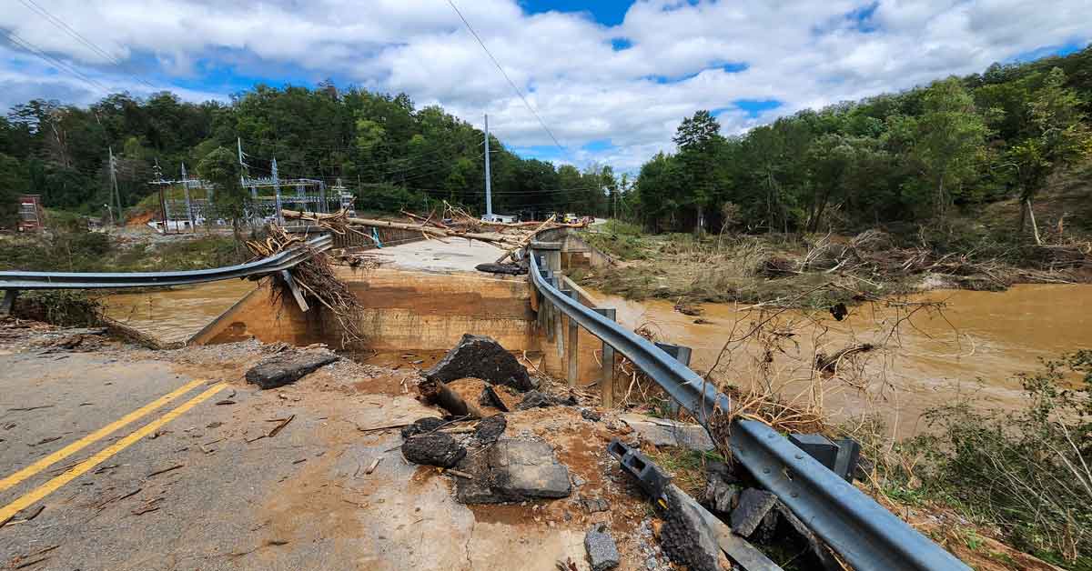 A road with metal barriers on either side and water running through the middle, the road is caved in and pieces of tree, asphalt are littered on the sides
