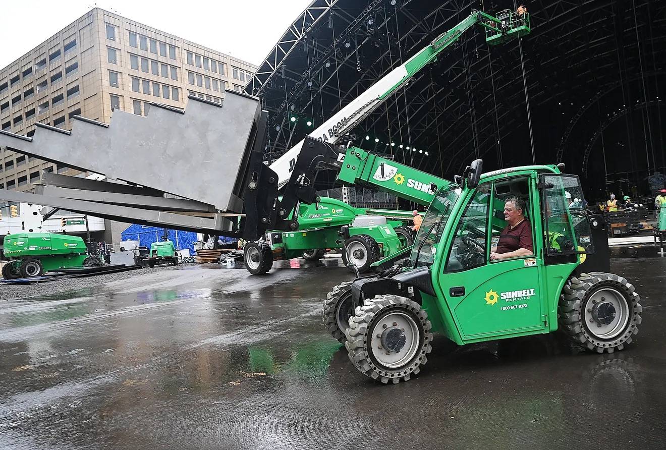 A green lift truck carring a set of metal risers away from a stadium building