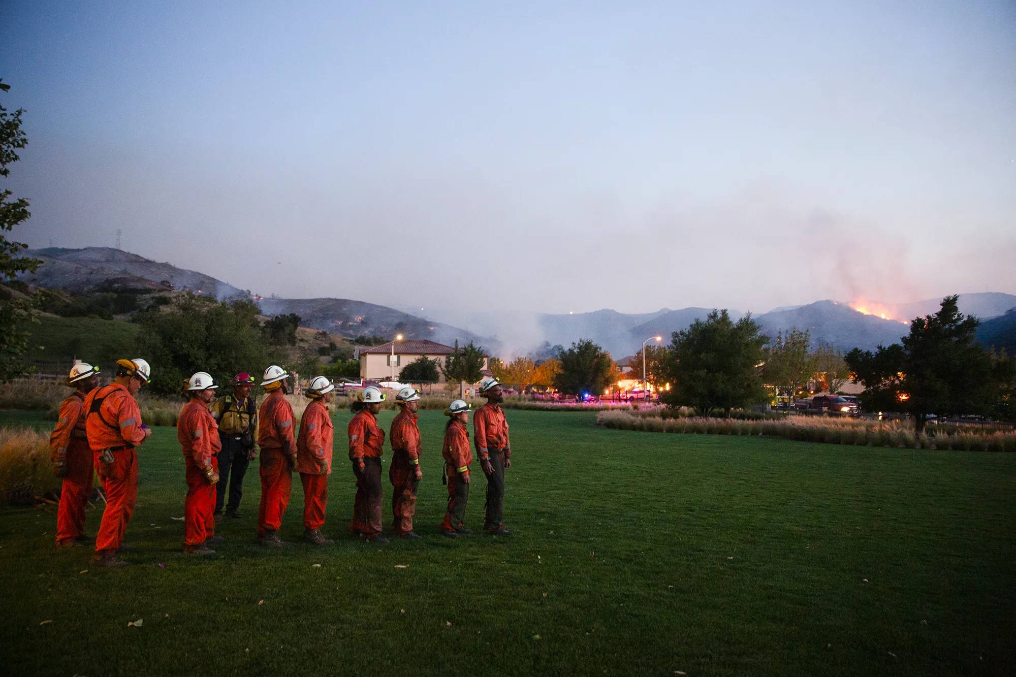 Firefighters in red suits and white helmets stand in a line on a grass field at dusk, with mountains burning in the background and a town with lit buildings in the middle distance.