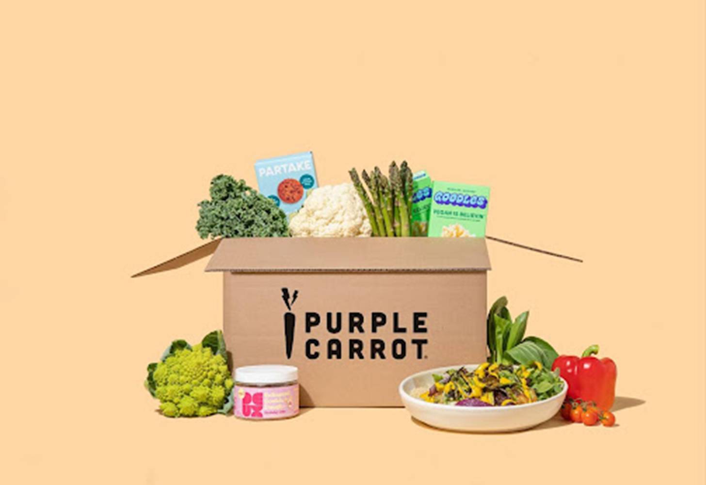 A brown Purple Carrot-branded box with vegetables coming out of it and other food items around it