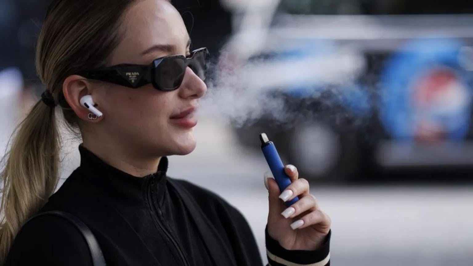 A person in a ponytail wearing a black shirt and black sunglasses holding a blue vape pen, they're blowing smoke out of their mouth