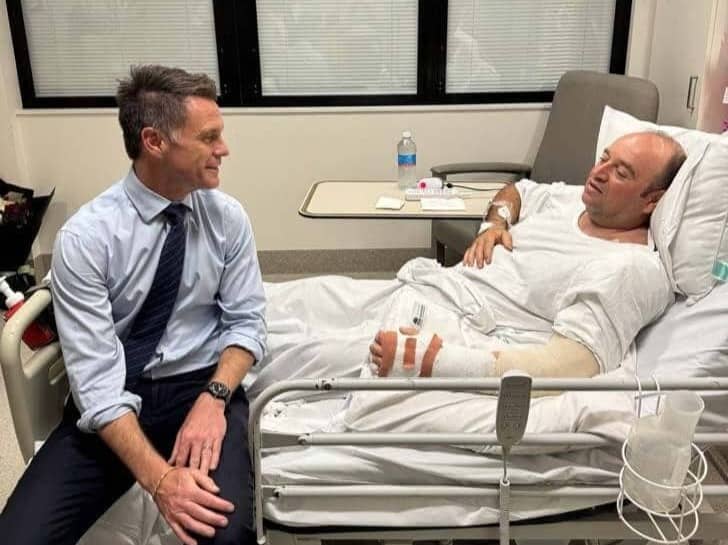 A person in a tie sitting on the edge of a hospital bed, there's a man in the bed with his arm in a cast