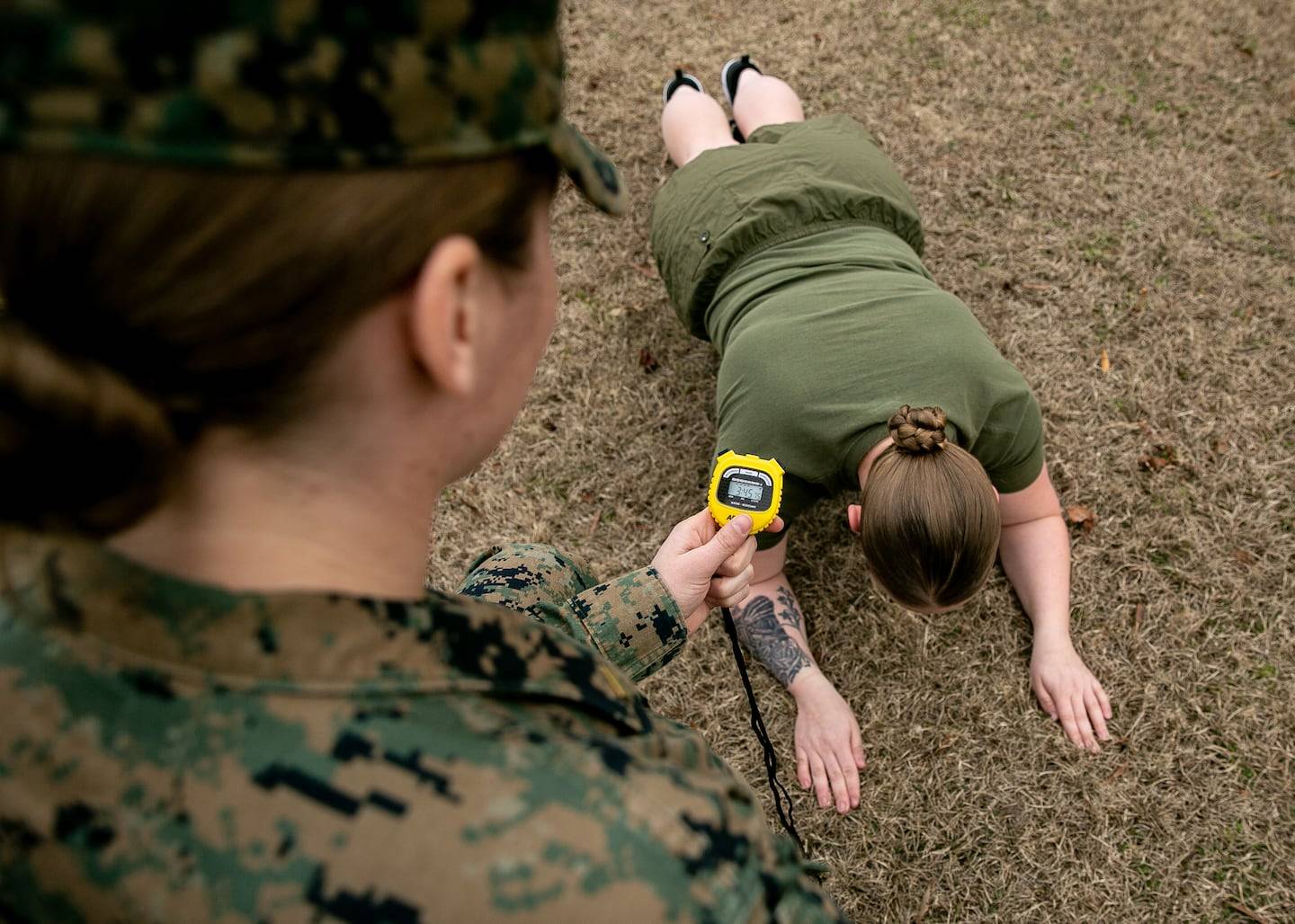A person in a green shirt lies on the ground holding a yellow digital watch on a cord while another person in camouflage clothing looks down at them.