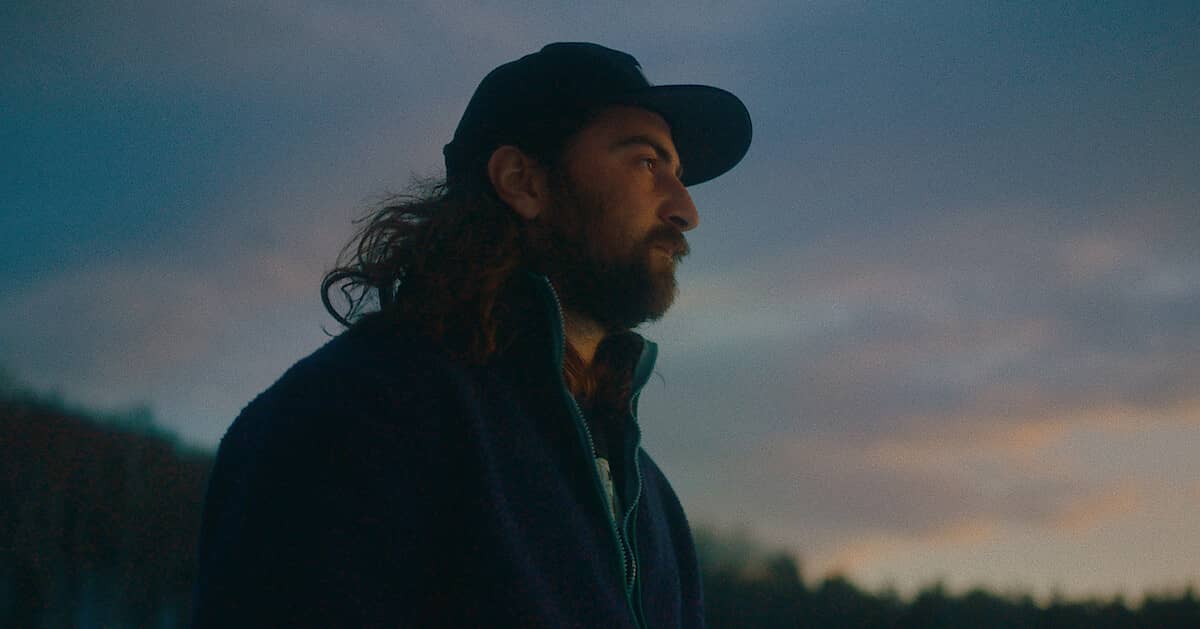 Noah Kahan looking into the distance at sunrise, he's wearing a jacket and baseball hat