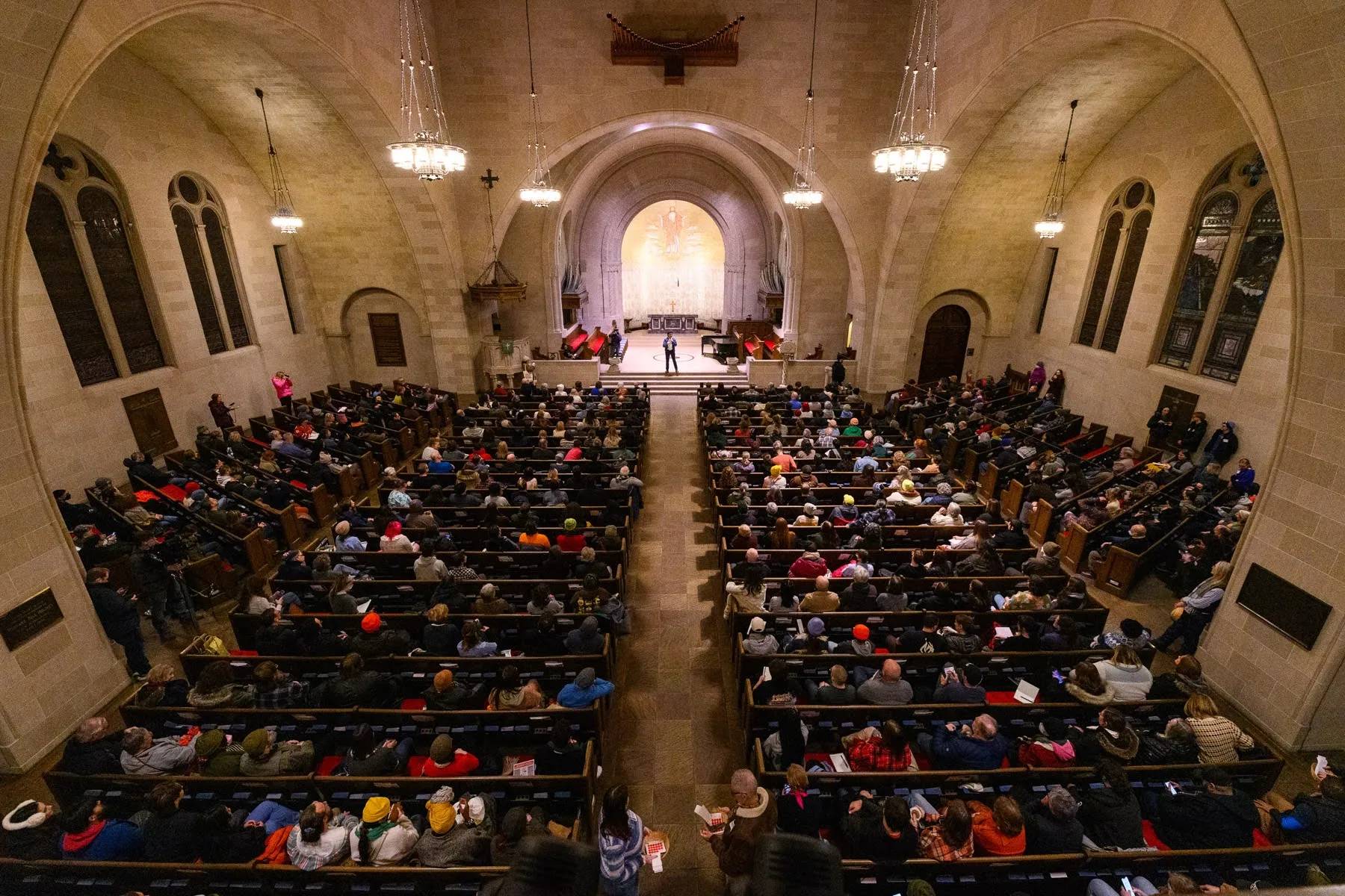 A church packed with people, a person stands at the front with a microphone